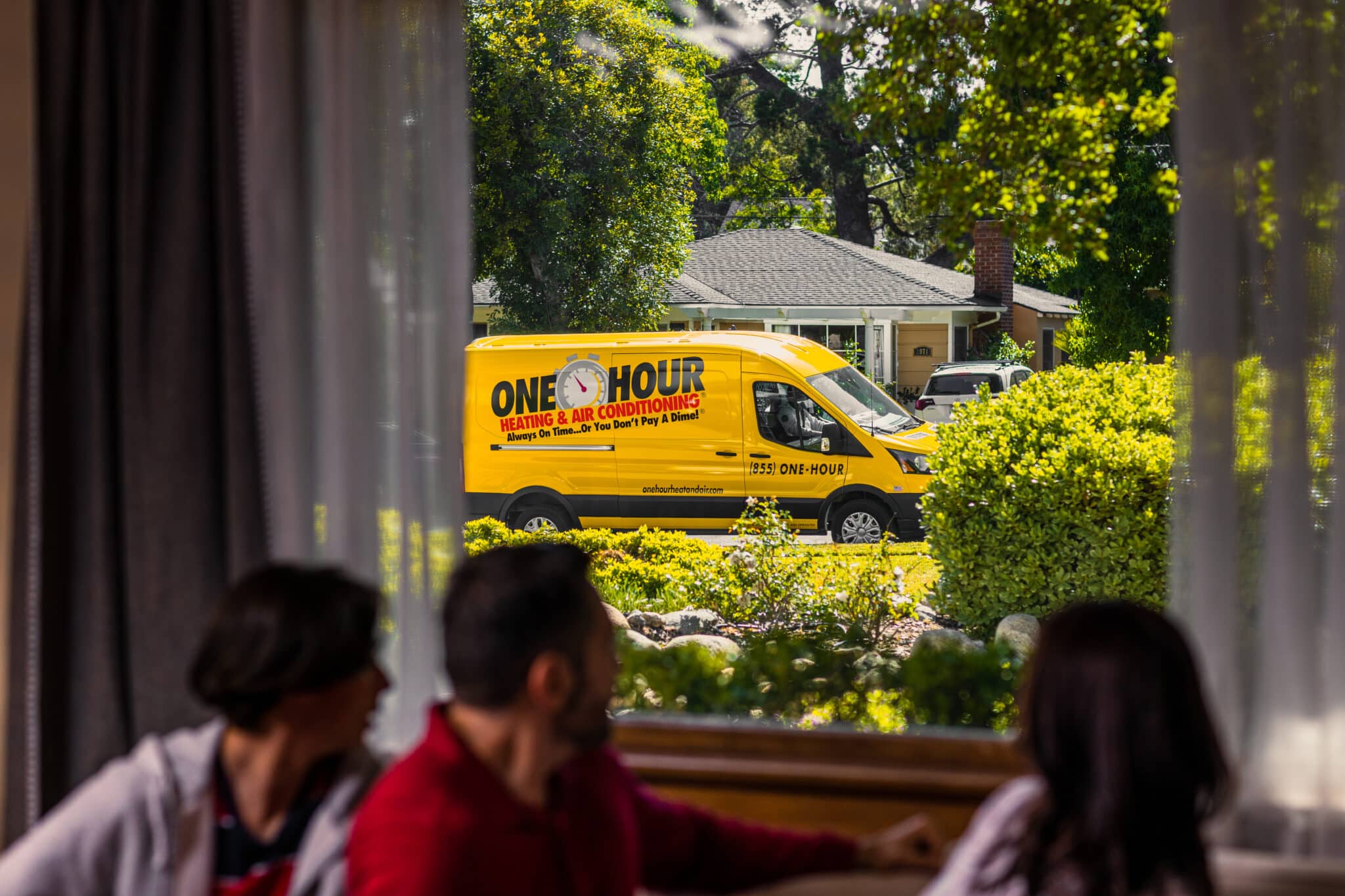 Family sitting on the couch inside, turned around looking out of the window at a One Hour Heating & AC van pulling up on the curb outside.